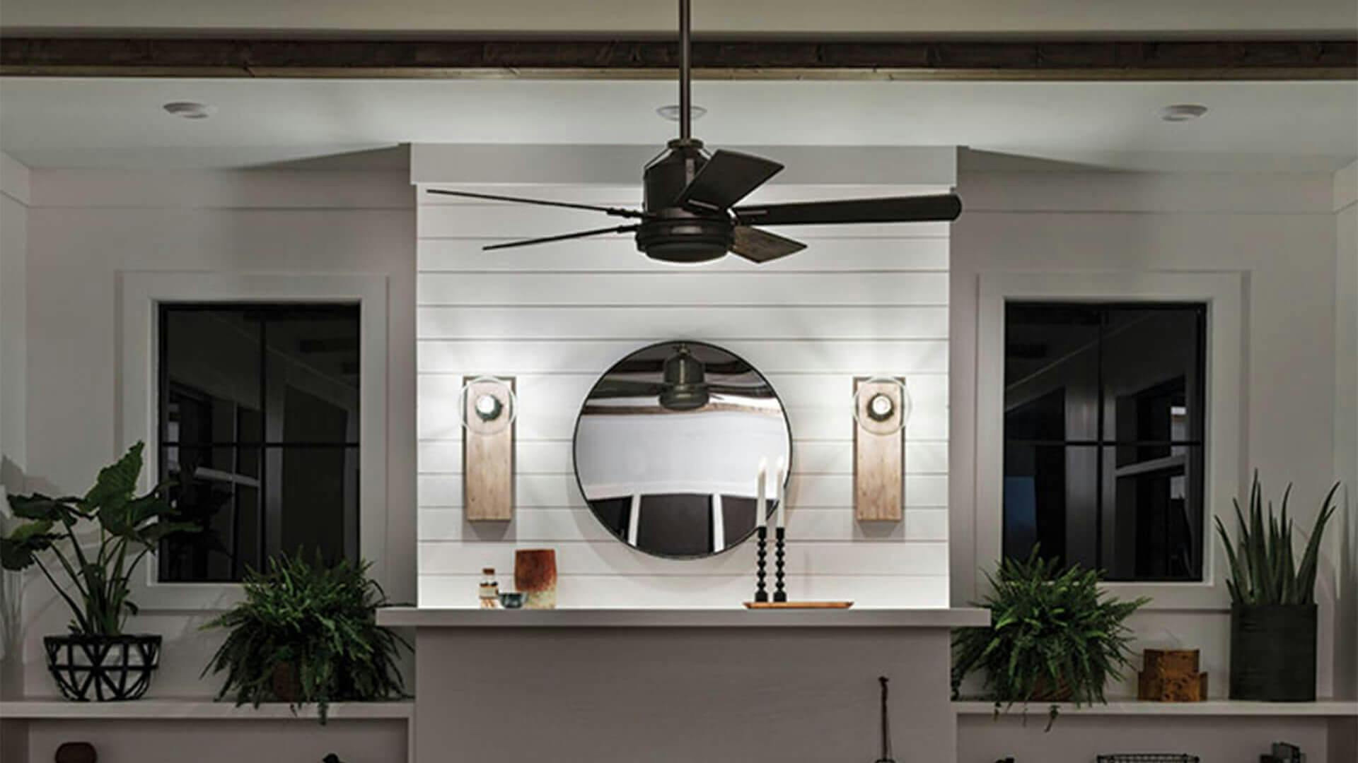 Farmhouse-style living room lit by two sconces centered above the fireplace on each side of a mirror while a ceiling fan hangs above